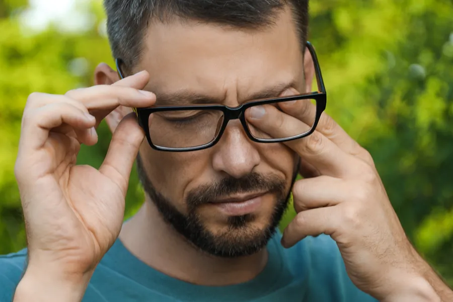 Allergic-Conjunctivitis-Clinic A man with a beard and glasses standing outside and wearing a blue t-shirt lifts his glasses and rubs his eyes due to allergy irritation. Get treatment for allergic conjunctivitis from Dr. Donna Diamond in Louisville/Boulder.