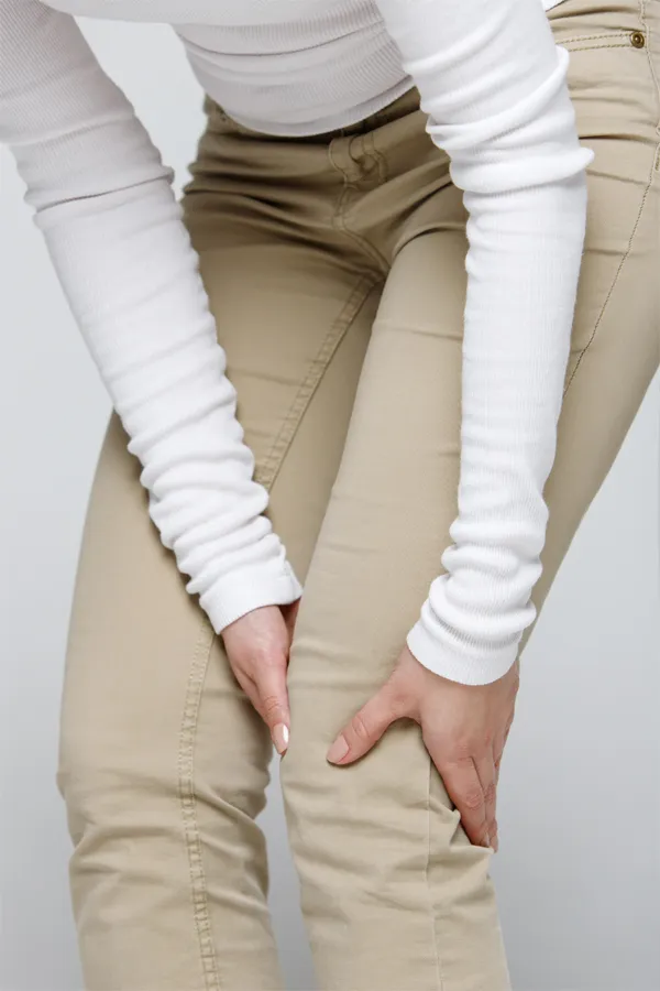 Arthritis-Treatment A woman wearing a white long-sleeve shirt and tan pants, clutching her knee, suffering from arthritis. Get arthritis treatment from Dr. Donna Diamond in Louisville/Boulder.