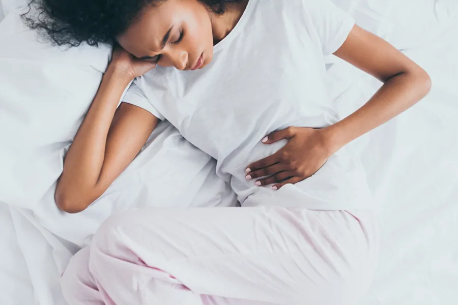 Crohns-Disease-Clinic A dark-skinned woman in white clothes lays on her bed clutching her middle before getting treatment for Crohn's Disease from Dr. Donna Diamond in Louisville/Boulder.