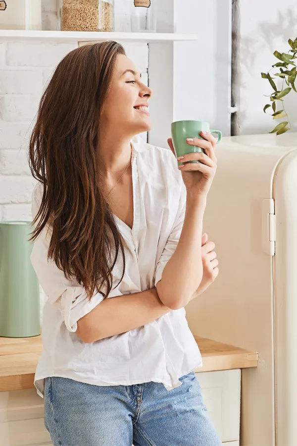 Crohns-Disease-Treatment A woman with a white blouse an jeans drinking tea in her kitchen, smiling after getting treatment for Crohn's Disease from Dr. Donna Diamond in Louisville/Boulder.
