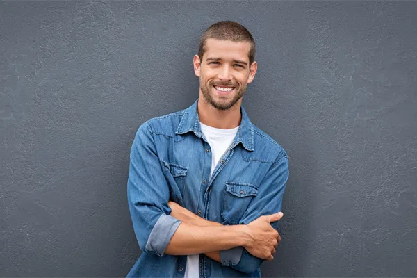 Hormones-for-Men-Doctor A man in a denim shirt stands smiling against a gray-blue wall, pleased with his testosterone hormone therapy from Dr. Donna Diamond in Louisville/Boulder.