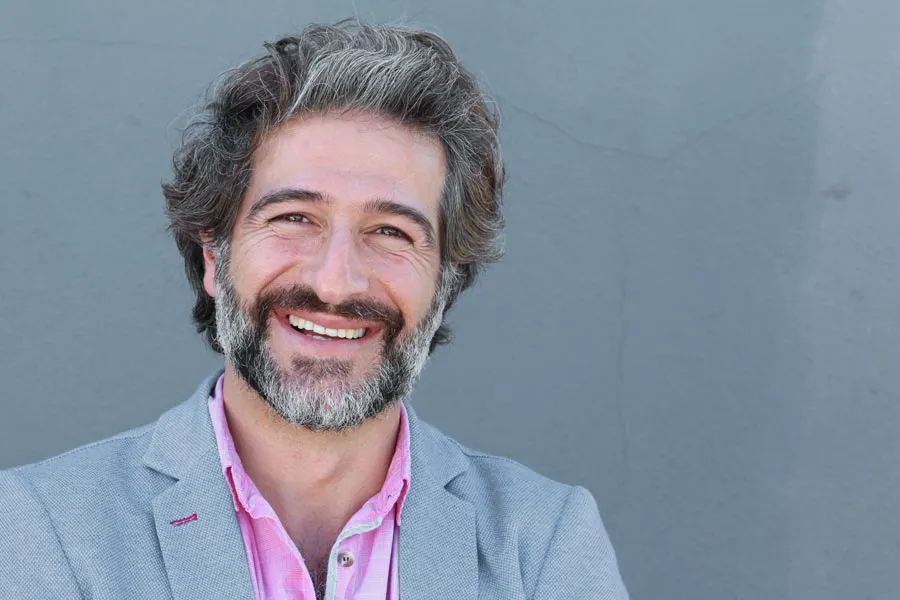 Hypogonadism-Clinic A smiling man with salt and pepper hair and beard wearing a pink shirt and gray blazer stands agains a gray wall. Get hypogonadism treatment from Dr. Donna Diamond in Louisville/Boulder.