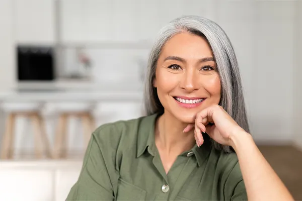 Menopause-Clinic An asian woman with long gray hair and a green shirt sitting and smiling on her couch, happy with her menopause care from [NAME} in Louisville/Boulder.