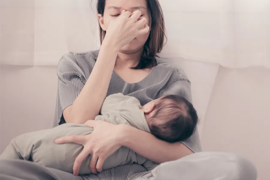 Postpartum-Hormone-Imbalance-Clinic A woman nursing an infant on couch rubs her eyes with exhaustion. Get treatment for postpartum hormone imbalance from Dr. Donna Diamond in Louisville/Boulder.